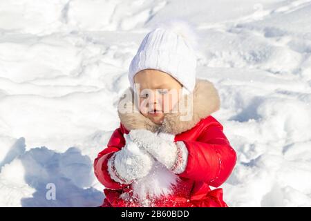 Activités hivernales pour les enfants. Une petite fille essaie de faire un boule de neige. Elle est habillée d'un manteau rouge vif avec un chapeau blanc, un foulard et des mitaines Banque D'Images