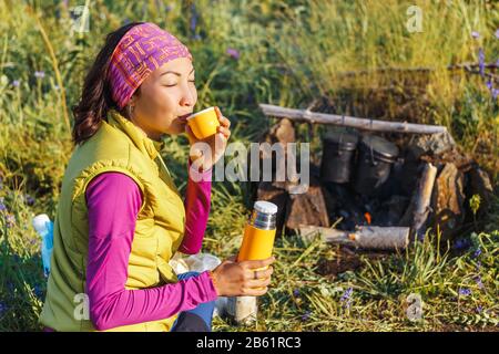 Femme voyageur tenant une bouteille thermos et une tasse de thé devant un feu de camping Banque D'Images