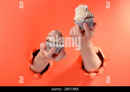 Les mains tenant deux cupcakes par des trous déchirés sur fond de papier rouge Banque D'Images