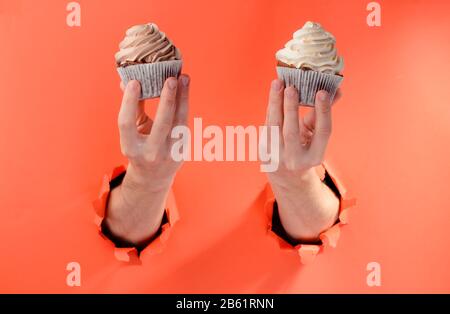 Les mains tenant deux cupcakes par des trous déchirés sur fond de papier rouge Banque D'Images