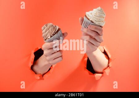 Les mains tenant deux cupcakes par des trous déchirés sur fond de papier rouge Banque D'Images