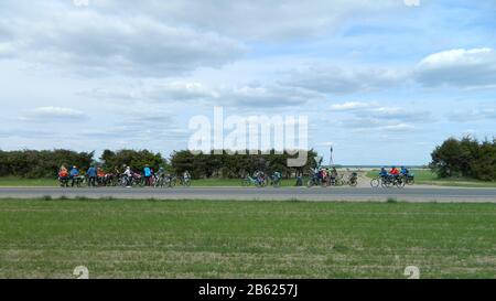 Un groupe de touristes sur les vélos se déplace le long d'une route asphaltée à travers la campagne contre un fond de buissons verts, herbe et un ciel nuageux. Banque D'Images