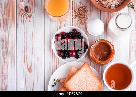 Délicieux petit déjeuner sain et copieux sur la table blanche. Œufs, flocons d'avoine, lait, thé, pain grillé à la banane, caramel salé maison, crêpes aux fruits, orange Banque D'Images