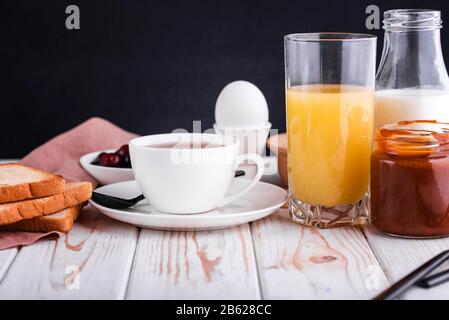 Délicieux petit déjeuner sain et copieux sur la table blanche. Œufs, flocons d'avoine, lait, thé, pain grillé à la banane, caramel salé maison, crêpes aux fruits, orange Banque D'Images