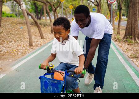 Sourire Heureux père africain américain aide son fils à apprendre le vélo dans le parc. Bonne Famille Bonding Loisirs Plein Air Concept De Sports Banque D'Images