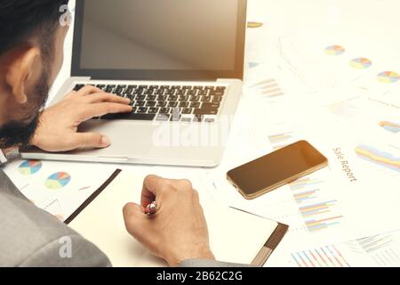 homme d'affaires assis écrire du texte note couverture noire sur le diagramme papier regardant ordinateur portable, il porte costume gris et utilise le stylo rouge et smartphone sur Banque D'Images