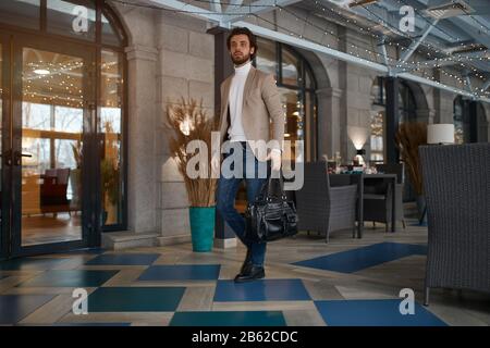 homme élégant en jean, veste beige, col roulé tenant un sac en cuir marchant à l'aéroport, allant à la salle d'attente, départ . l'homme arrive à la a. Banque D'Images