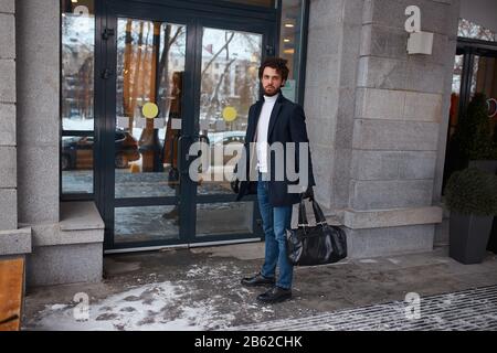 l'homme est prêt pour un voyage d'affaires, élégant homme est en train de regarder l'appareil photo, tout en se tenant près de la porte du bâtiment, pleine photo Banque D'Images