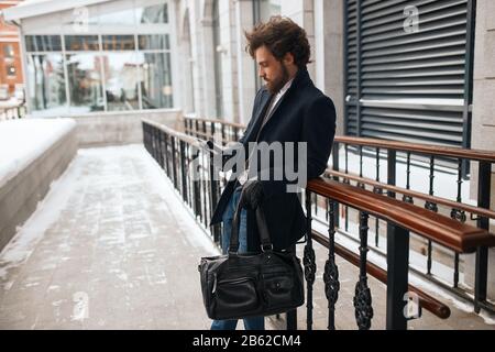 beau homme qui tient le smartphone , sac ayant un repos à l'extérieur, en attendant une petite amie, date, vue rapprochée photo, crime, homme en attente Banque D'Images