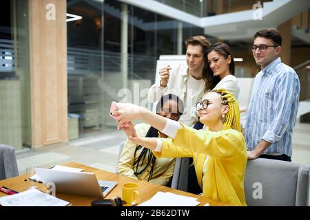 Joyeux et joyeux collègues qui se font selfie avec le téléphone de vente. Intérieur de bureau, papeterie, papier, ordinateur portable sur la table, vue latérale rapprochée photo Banque D'Images