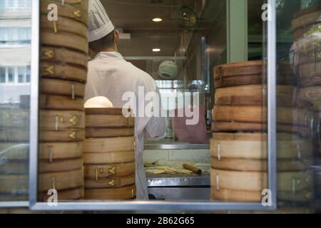 Chef chinois debout dans une cuisine de boulangerie, entouré de boulettes de bambou boîtes, vu par une fenêtre. Banque D'Images