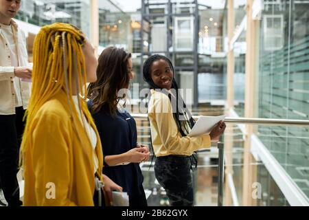 afro femme souriante avec des dreads dans un élégant chemisier jaune tenant des doculants dans les mains regardant en arrière, à ses collègues dans la salle de bureau Banque D'Images