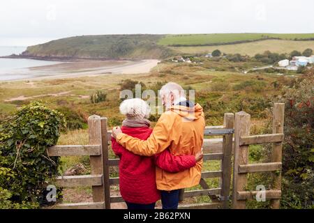 Vue Arrière D'Un Couple Senior Actif Regardant Par-Dessus La Porte Pendant Qu'Ils Marchent Le Long Du Sentier Côtier À L'Automne Banque D'Images