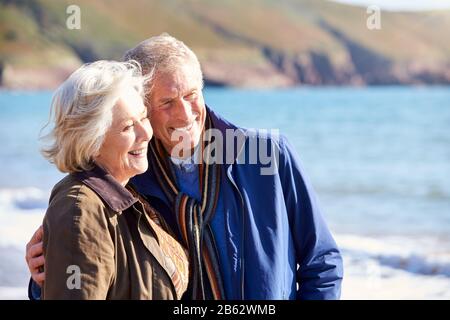 Un Couple Senior Affectueux S'Embrassant LorsQu'Ils Marchent Le Long De Shoreline Of Beach By Waves Banque D'Images