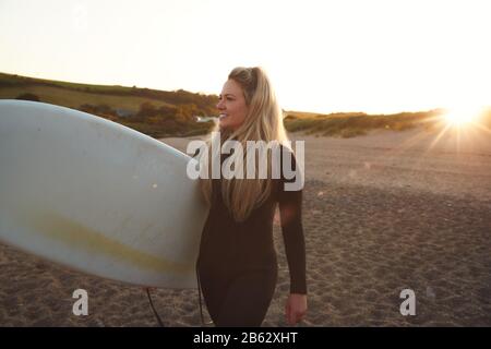 Femme Portant Wetsuit Holding Surfboard En Profitant De La Staycation De Surf Sur La Plage Comme Sun Sets Banque D'Images