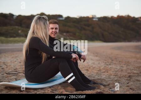 Couple Portant Des Combinaisons Sur Le Surf Staycation Assis Sur Surfboard Donnant Sur La Mer À Waves Banque D'Images
