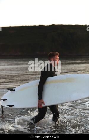 Homme Portant Des Wetsuit Transportant Surfboard LorsQu'Il Sort De La Mer Banque D'Images
