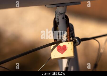 Cadenas blanc avec coeur rouge sur une clôture. Symbole de l'amour, de la relation , cadeau de Saint Valentin. Fond orange brun foncé flou Banque D'Images