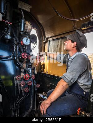 Jeune homme faisant du bénévolat comme pompier de moteur à vapeur et ingénieur assis dans le moteur de locomotive à vapeur regardant la fenêtre avec la main sur les commandes. Banque D'Images