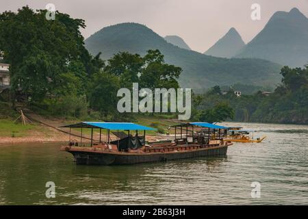 Guilin, Chine - 10 Mai 2010 : Le Long De La Rivière Li. Navire de transport en vrac métallique avec feuillage vert sur la rive et les pics de montagne karst sous le ciel gris comme backdro Banque D'Images