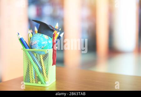 Éducation apprentissage et retour à l'école avec un chapeau de graduation sur un étui crayon sur la table dans la bibliothèque contexte / chapeau de graduation sur terre globe mode Banque D'Images