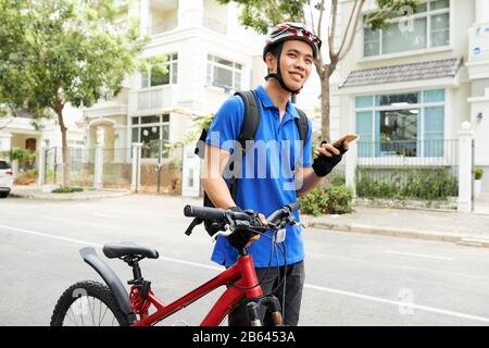 Un jeune homme souriant et souriant à la livraison vietnamienne se tenant à côté de sa bicyclette avec un smartphone à portée de main Banque D'Images