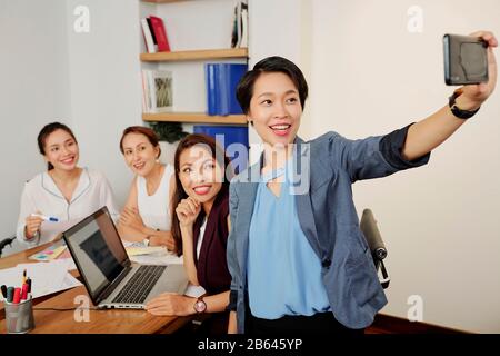 Bonne jeune fille d'affaires qui prend selfie avec ses collègues lors de la réunion Banque D'Images
