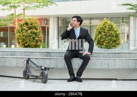 Jeune entrepreneur assis à l'extérieur à côté de son scooter électrique et appelant au téléphone Banque D'Images