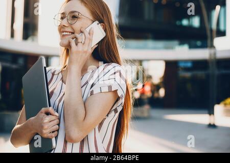 Une femme d'affaires caucasienne aux cheveux rouges est en possession d'un ordinateur portable lors de la marche et de la conversation au téléphone Banque D'Images