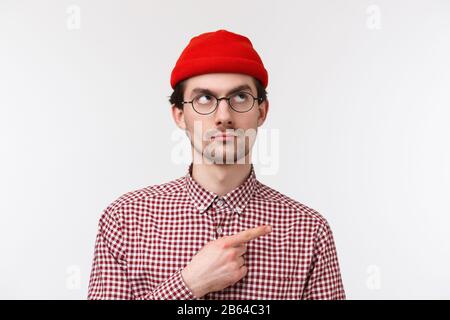 Sceptique bon look jeune hipster gars en bonnet rouge et vérifier chemise, lunettes, peeking douteux, regardant pointant vers le coin supérieur droit avec sérieux Banque D'Images