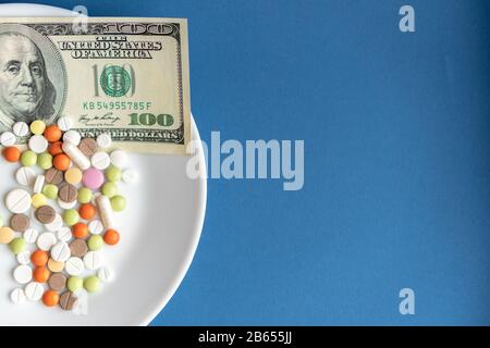 Les médicaments coûtent de l'argent. Médicaments et dollars.plaque blanche avec pilules et capsules multicolores et une facture de 100 dollars sur fond bleu classique. Pilules Banque D'Images