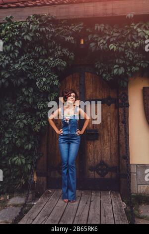 Portrait d'une belle femme portant une combinaison denim debout contre une ancienne porte en bois à cadre ivy d'un château. Ton magique Banque D'Images