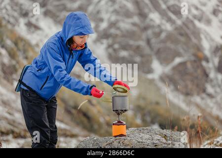 Femme bouillante sur un jet de gaz portable de camping près de la tente de randonnée dans les montagnes enneigées de la fin de l'automne Banque D'Images