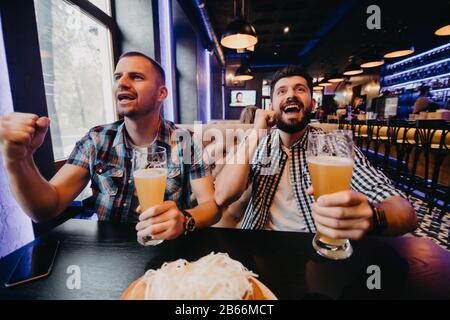 Deux jeunes hommes enthousiastes et gaimés qui regardent le match sur un ordinateur portable et soutiennent leur équipe assis au bar Banque D'Images