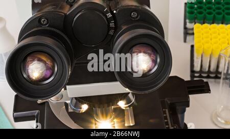 Lentille oculaire de microscope médical de laboratoire avec miroir d'un visage d'homme dans un masque chirurgical blanc. Dispositif de diagnostic optique et tubes à essai en plastique dans les portoirs. Banque D'Images