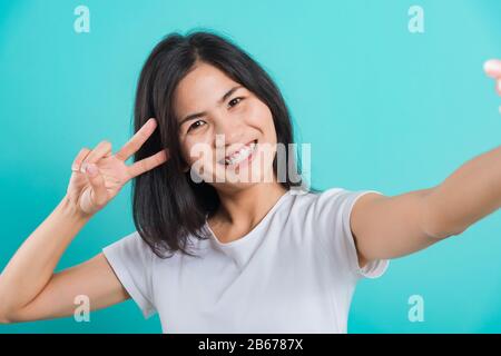 Portrait Happy Asian belle jeune femme sourire blanc dents debout porter un t-shirt blanc, Elle fait selfie avant appareil photo smartphone montrant V-Sign, loo Banque D'Images