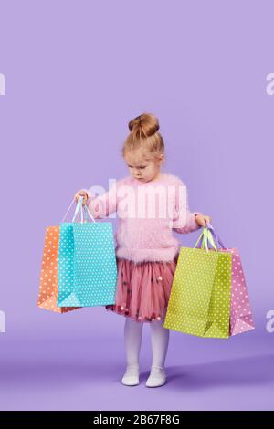 Portrait d'une heureuse petite fille d'enfant debout sur un fond mauve tenant des sacs de shopping, paquet. Regarder l'appareil photo. Banque D'Images