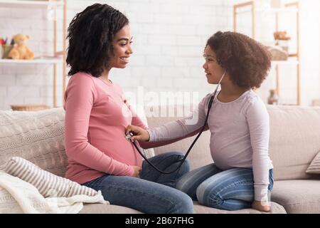 Petite Fille Jouant Avec La Mère À La Maison, Écoutant La Ceinture Enceinte De Maman Banque D'Images