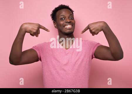 Jeune homme africain positif souriant et se pointant avec index doigts. Beau jeune homme portant une chemise rose gestuelle et regardant l'appareil photo. Banque D'Images