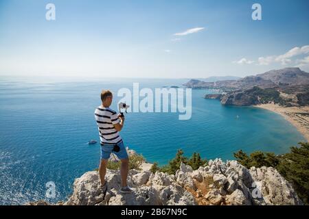 Homme filmant sur la caméra en haut de la montagne. Randonnée touristique et prise de photos. Vue panoramique sur la plage de Tsampika, les montagnes et la mer bleue, Rhodes Banque D'Images