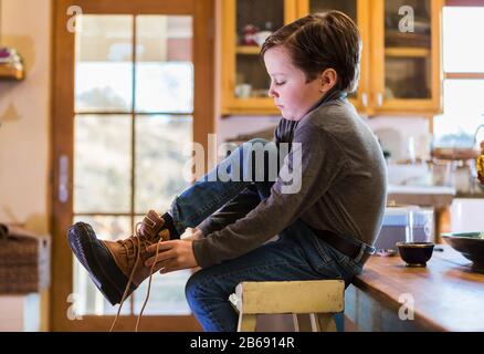 Un garçon de six ans qui met ses bottes sur place, assis sur un haut tabouret. Banque D'Images