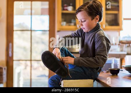 Un garçon de six ans qui met ses bottes sur place, assis sur un haut tabouret. Banque D'Images