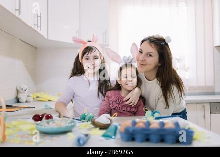 Les enfants portent des oreilles de lapin posant avec leur mère. Sœurs et maman souriant dans la cuisine et regardant l'appareil photo tout en se préparant pour les vacances de Pâques. Banque D'Images