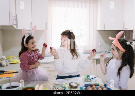 Mère et filles qui détiennent des œufs rouges peints. Joyeux maman et mignonne petites filles portant un lapin tenant des œufs de Pâques rouges dans la cuisine avec un accessoire de peinture Banque D'Images