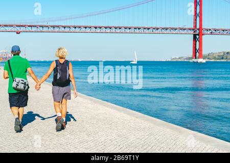 Heureux couple à la retraite de femme et d'homme avec sac pack marchant main dans la main, explorer Lisbonne et profiter de la vue sur le pont rouge Ponte 25 de Abril. Suspensi Banque D'Images