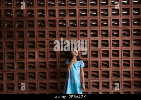 petite fille se tient avec son geste de main tenant son menton devant un mur avec des briques avec un motif creux Banque D'Images