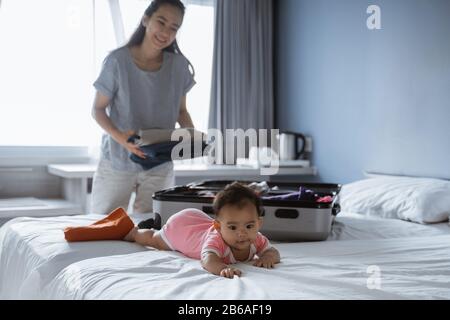 un bébé mignon repose sur le lit quand sa mère met ses vêtements dans une valise avant de partir en vacances Banque D'Images