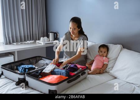 bébé se trouve à côté de la mère préparant des vêtements à mettre dans la valise Banque D'Images