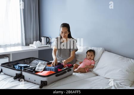 le bébé mignon se trouve à côté de la mère préparant des vêtements à mettre dans la valise Banque D'Images