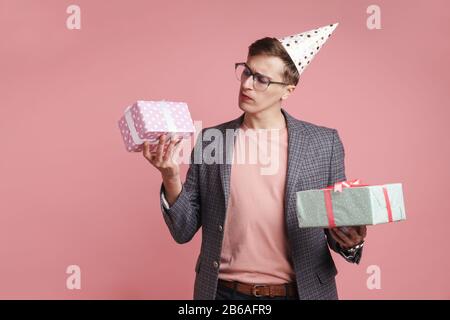 Image d'un jeune homme sérieux de pensée dans des verres tenant anniversaire cadeau boîtes isolées sur fond de mur rose. Banque D'Images